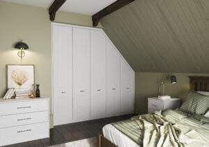 A cozy attic bedroom with sloped wooden ceiling and exposed beams. It features a neatly made bed with striped bedding, a white dresser, a nightstand with a lamp, and built-in white closets. The walls are painted a soft green.