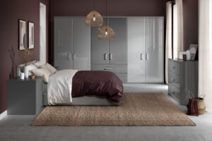 A modern bedroom with a large bed covered in beige and burgundy bedding. Grey wardrobes line the back wall, and two wicker pendant lights hang above. A woven rug lies on the wooden floor, and light pours through a window with sheer white curtains.