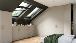 A modern attic bedroom with skylights, built-in cream wardrobes, and wooden parquet flooring. A neatly made bed with a green throw is in the foreground, and theres a small counter with books, plants, and decor items under the windows.