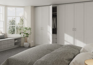 A bright bedroom with a cozy bed, gray blanket, and several pillows. Built-in white wardrobes with open doors reveal hanging clothes. Large windows provide natural light. A small plant and a window seat with drawers complement the space.