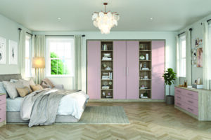 A spacious bedroom with a herringbone wood floor and a light color palette. It features a neatly made bed, pink wardrobe with shelves displaying decor, a plant, and art on the walls. A large window allows natural light to fill the room.